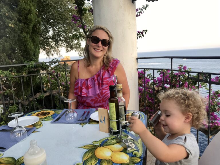 A smiling woman and a young child at a terrace table with a lemon-print cloth, above the sea on the Amalfi Coast