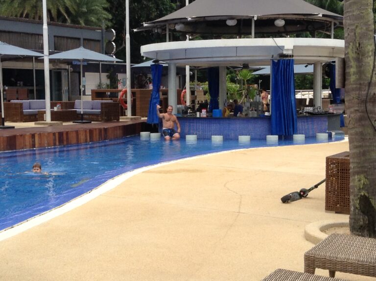 A swim-up bar with blue curtains beside a curving pool on Sentosa Island, with a man waving from a bar stool