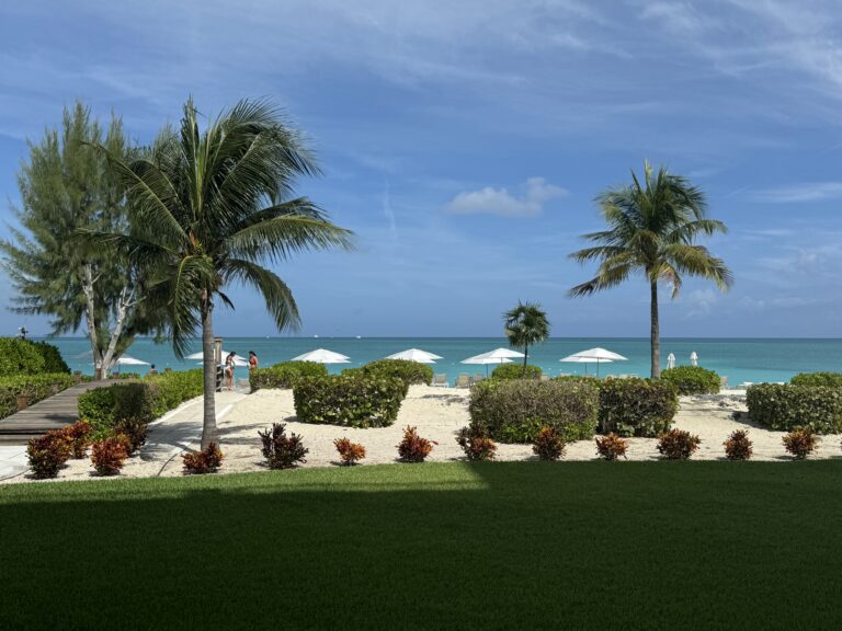 Palm trees and white parasols on the beach at The Venetian on Grace Bay, with the turquoise sea beyond a green lawn