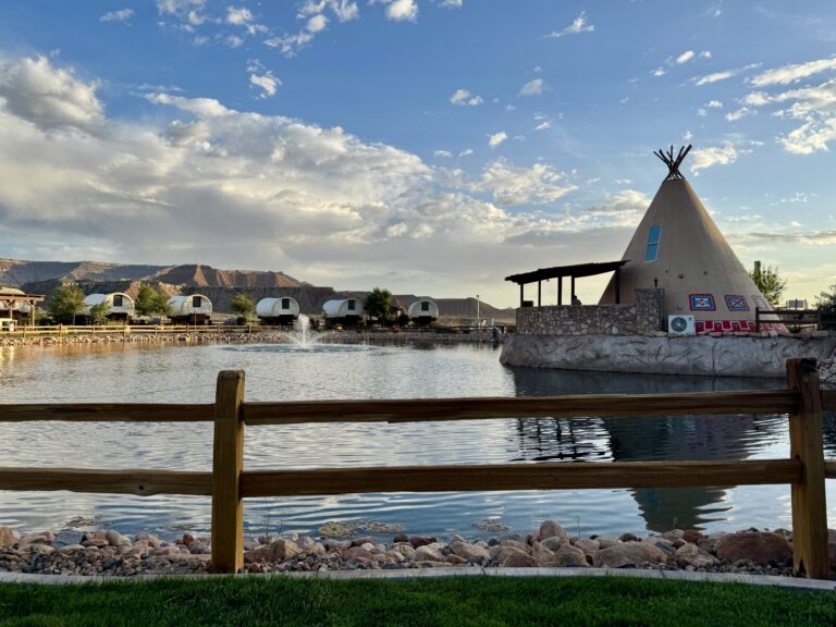 A teepee and covered wagons around a pond with a fountain at Zion White Bison Resort, with desert mesas in the distance