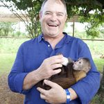 A laughing man in a blue shirt bottle-feeds a small monkey cradled in his arms under a leafy tree