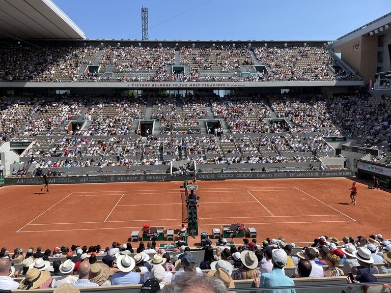 Packed stands around a red clay court during a French Open match, seen from behind spectators in sun hats