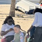 Two women braid a young girl's hair with colourful extensions on a sandy beach in the Dominican Republic