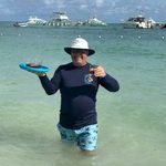A man in a sun hat stands in shallow turquoise water holding drinks on a blue frisbee, with boats moored behind