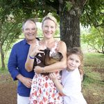 A family posing under a tree while a woman in a floral dress bottle-feeds a small monkey in her arms