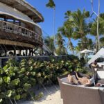 A multi-level wooden beach restaurant with a thatched roof, rattan loungers and tall palm trees in Punta Cana