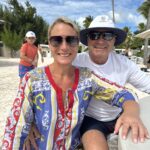 A smiling woman in a colourful kaftan sits with a man in a sun hat on a sandy beach lined with palm trees