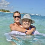 A smiling woman hugs a man in a bucket hat in the shallow turquoise sea off Punta Cana, with boats in the distance