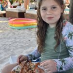 A young girl on a sandy beach holds a plate of grilled lobster, with sun loungers and umbrellas behind her