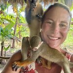 A smiling woman takes a selfie as two small monkeys climb over her shoulders under a thatched roof