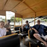 A young girl and three adults sit on black bench seats in an open-air tour vehicle, with palm trees outside