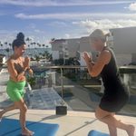 Two women doing a yoga pose on a balcony with blue mats, overlooking palm trees and the sea in Punta Cana