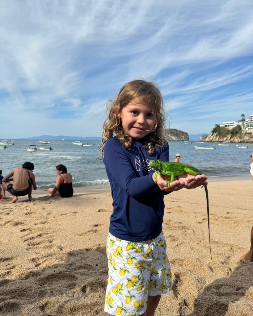 A young child holds a bright green iguana on a beach near Puerto Vallarta, with small boats in the bay behind