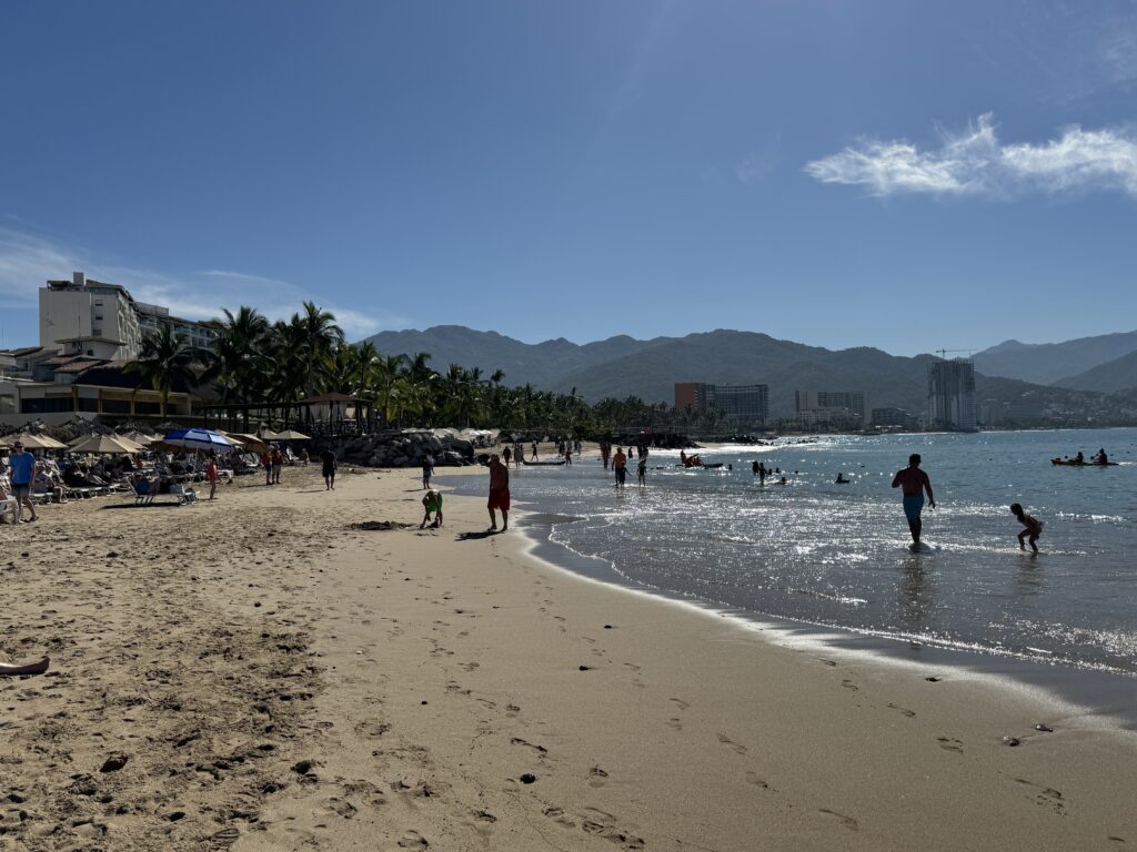 Sunny beach in Puerto Vallarta with people paddling in the sea, palm trees, hotels and green mountains behind