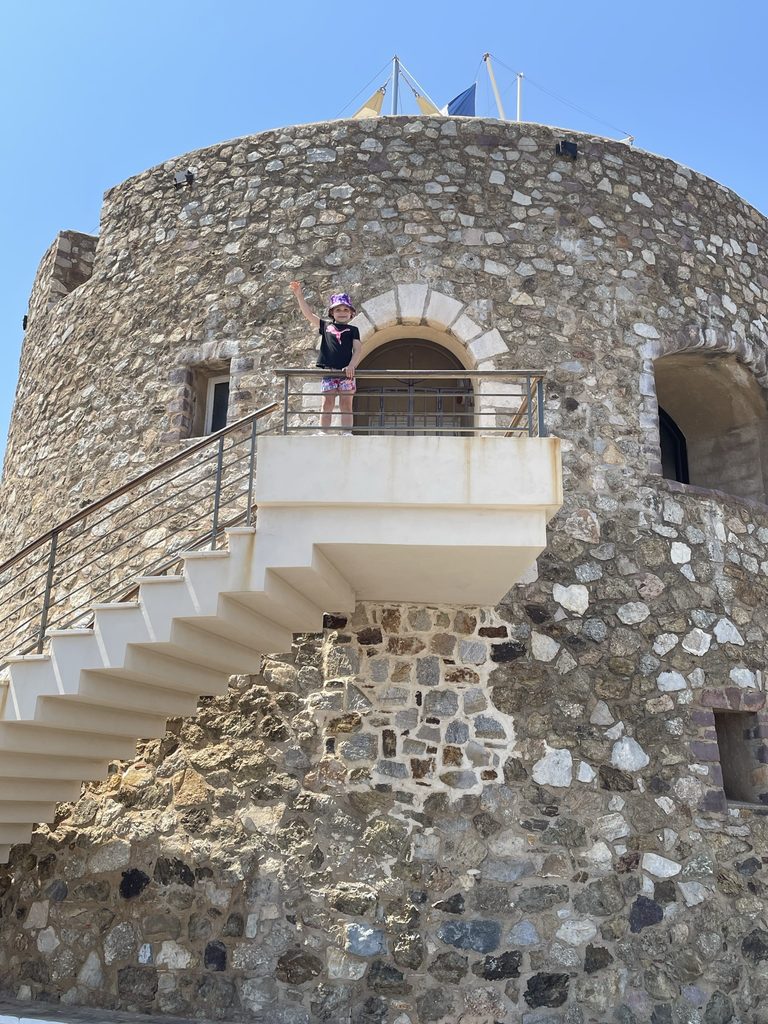 A young child in a purple sun hat waving from the balcony of a round stone tower with a white staircase