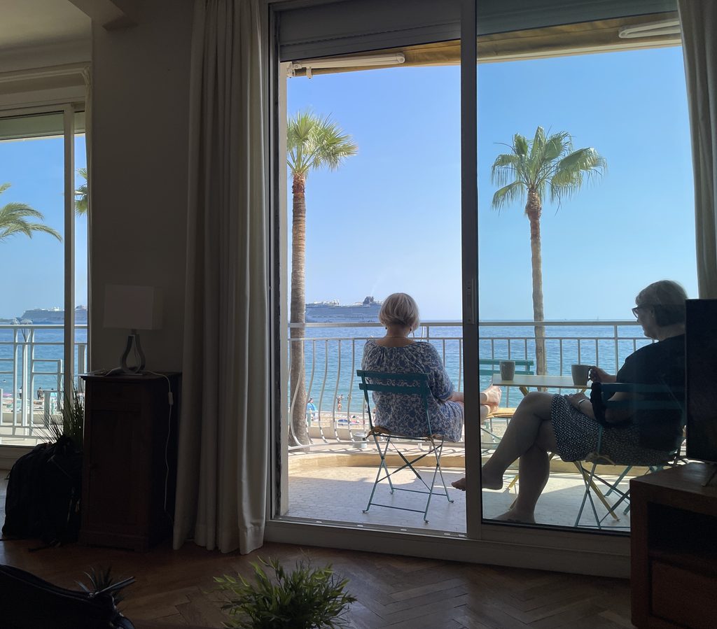 Two women relaxing with mugs on a sunny balcony overlooking palm trees, the sea and a cruise ship