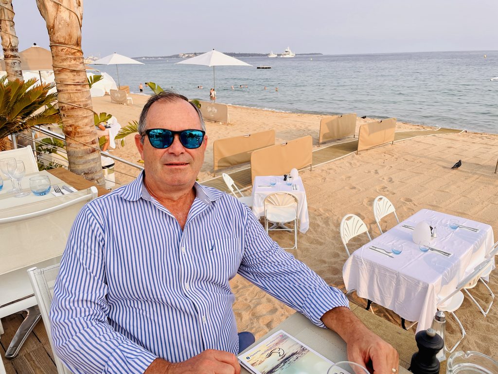 A man in a striped shirt at a French Riviera beach restaurant, with white-clothed tables, parasols and yachts behind